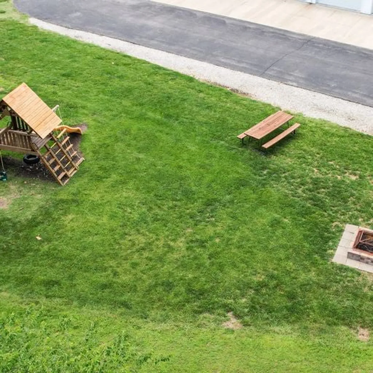 An inviting exterior space with a playground structure, picnic table, and a central fire pit, set against a backdrop of open sky and greenery. The setup encourages family gatherings, outdoor dining, and evenings by the fire.