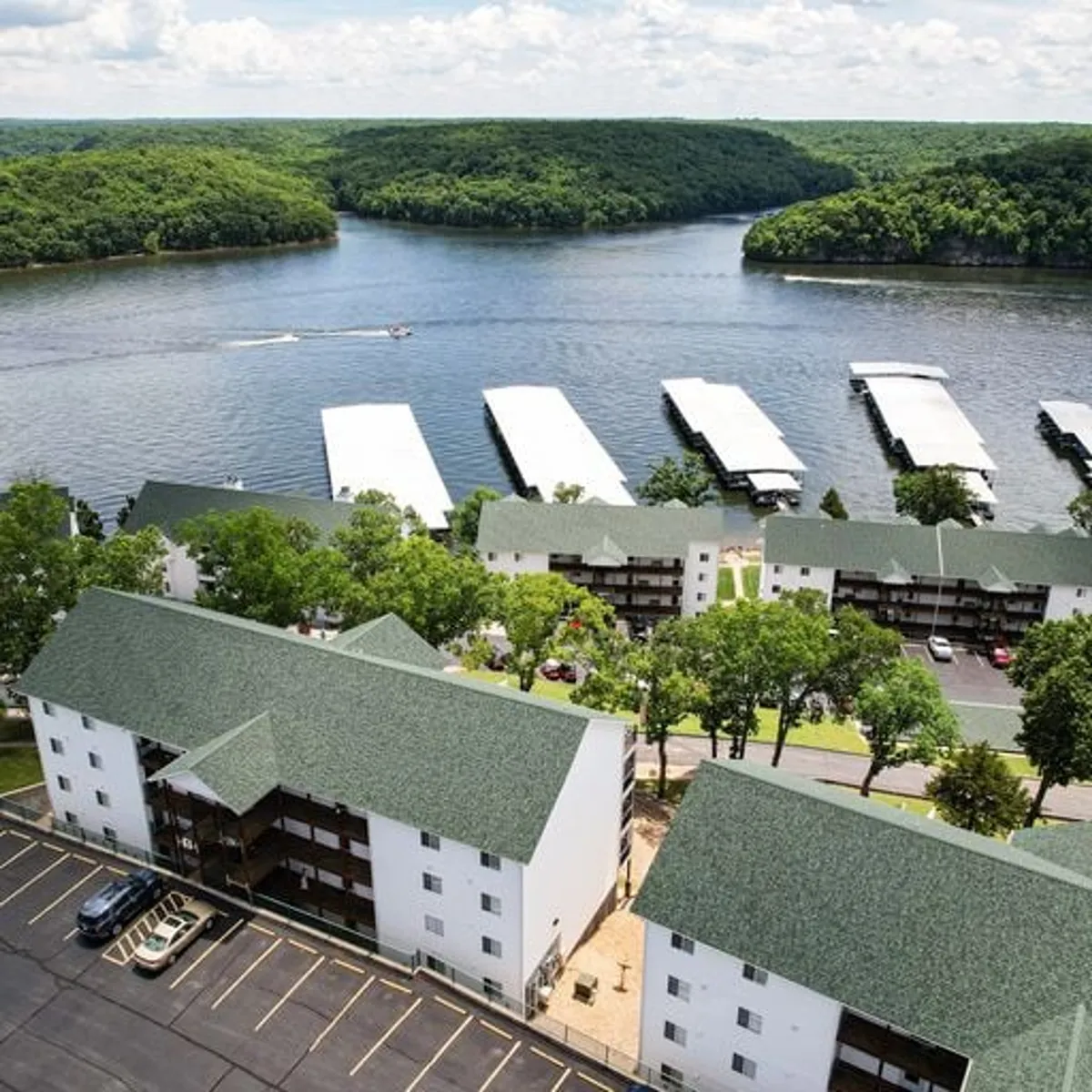 A tranquil lakeside scene featuring a calm lake bordered by green hills, with boathouses and apartments visible along the shore. The expansive shoreline hints at areas for picnicking and fishing, while the overall setting suggests easy access to outdoor adventures and relaxation.