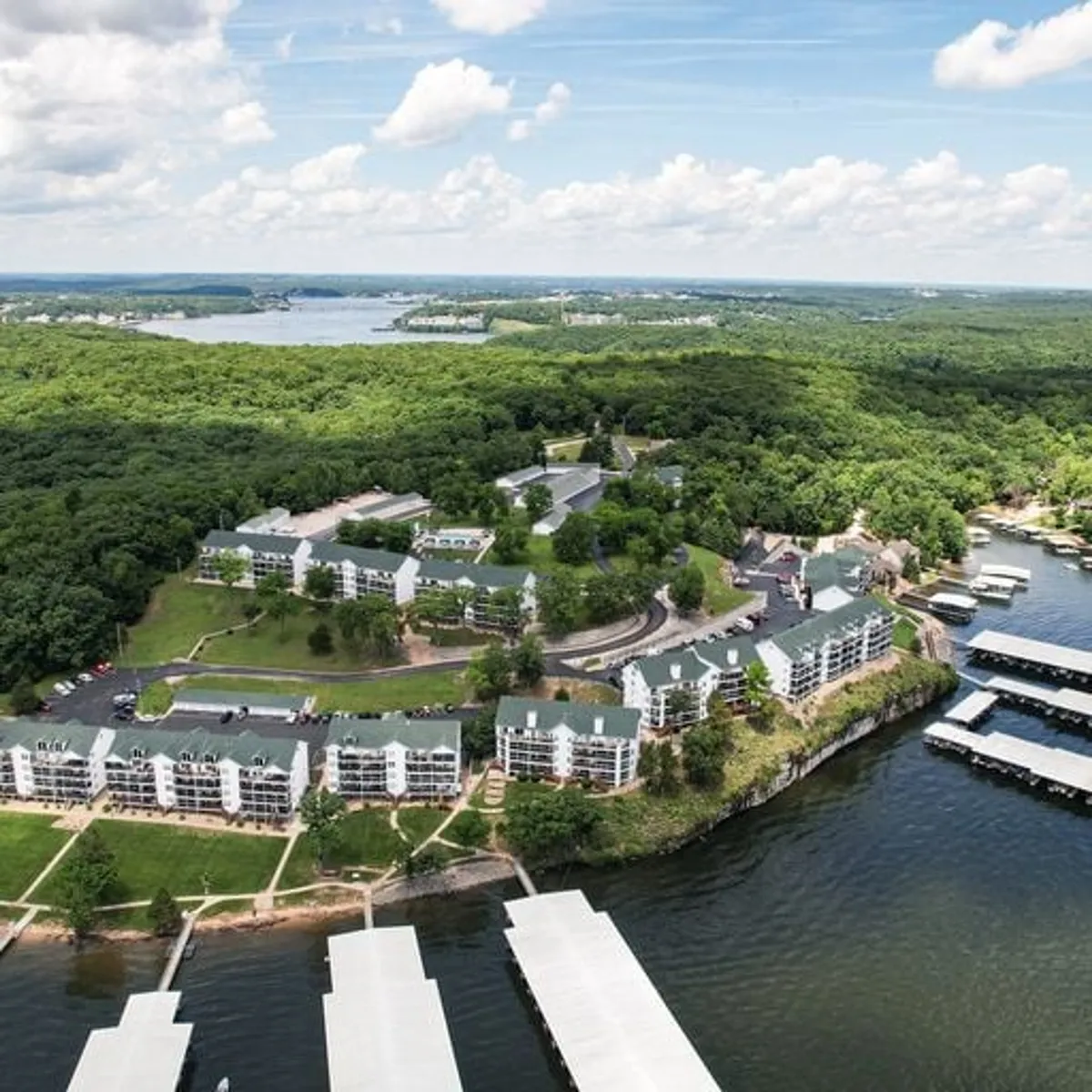A scenic lake vista featuring the broad sweep of the water, a wooded area along the shore, a marina with docked boats, and residential buildings nestled in the landscape. The calm water reflects the sky, and the perspective hints at activities like boating, fishing, and simply enjoying the outdoors.