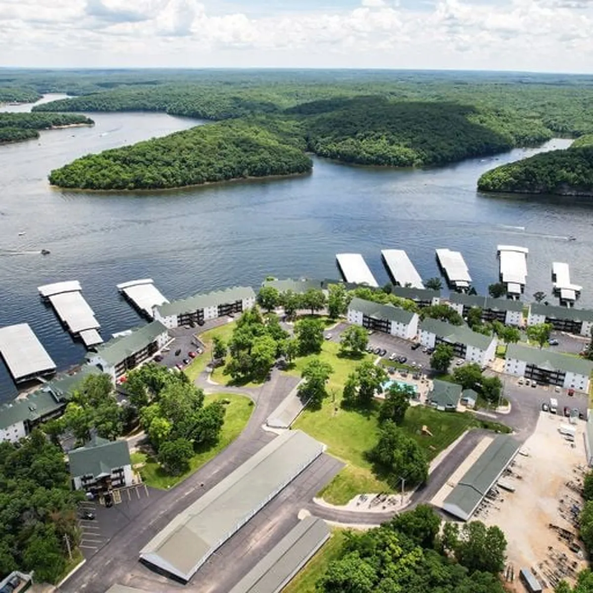 This exterior image captures a panoramic view of the lake, marina, green spaces, a pool area, and surrounding buildings. The photo blends natural beauty with recreational amenities, showing the variety of waterfront experiences available at Lake of the Ozarks.