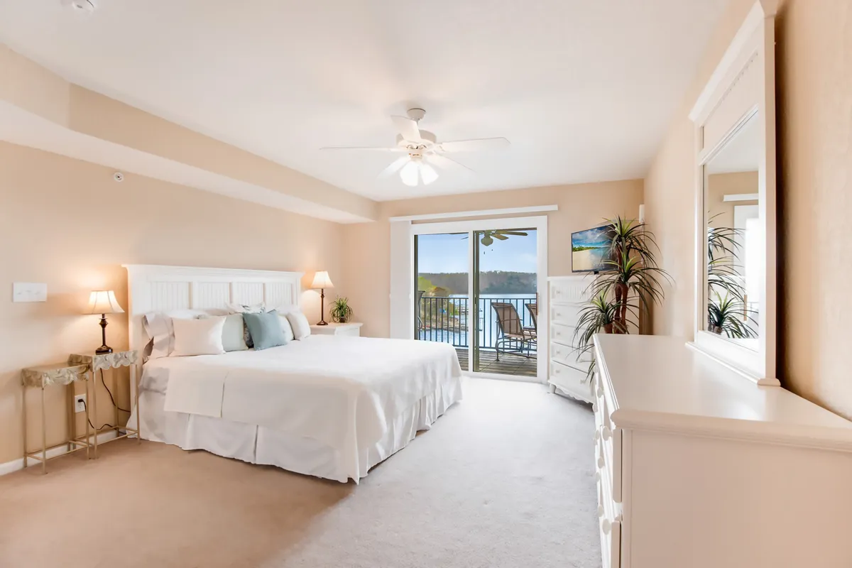 The master bedroom features a king-sized bed positioned to face balcony doors that open to expansive lake views. Decorative plants and a dresser with a mirror create a serene, uncluttered atmosphere, while natural light floods the space.