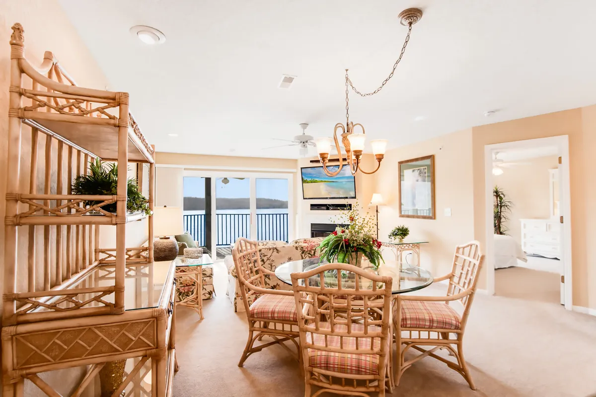 A bright dining area featuring a table with rattan chairs, illuminated by a chandelier and framed by large windows that offer picturesque views. Decorative shelves add warmth, and the space feels designed for gathering, sharing meals, and enjoying the surrounding scenery.