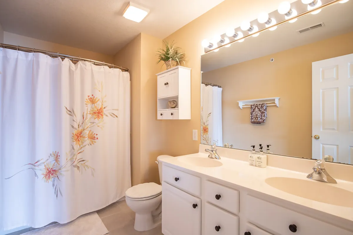 A calming bathroom space with a stylish vanity, large mirror, decorative shower curtain, and thoughtful accents on a shelf. The lighting and decor combine for a soothing, modern atmosphere ideal for daily routines.