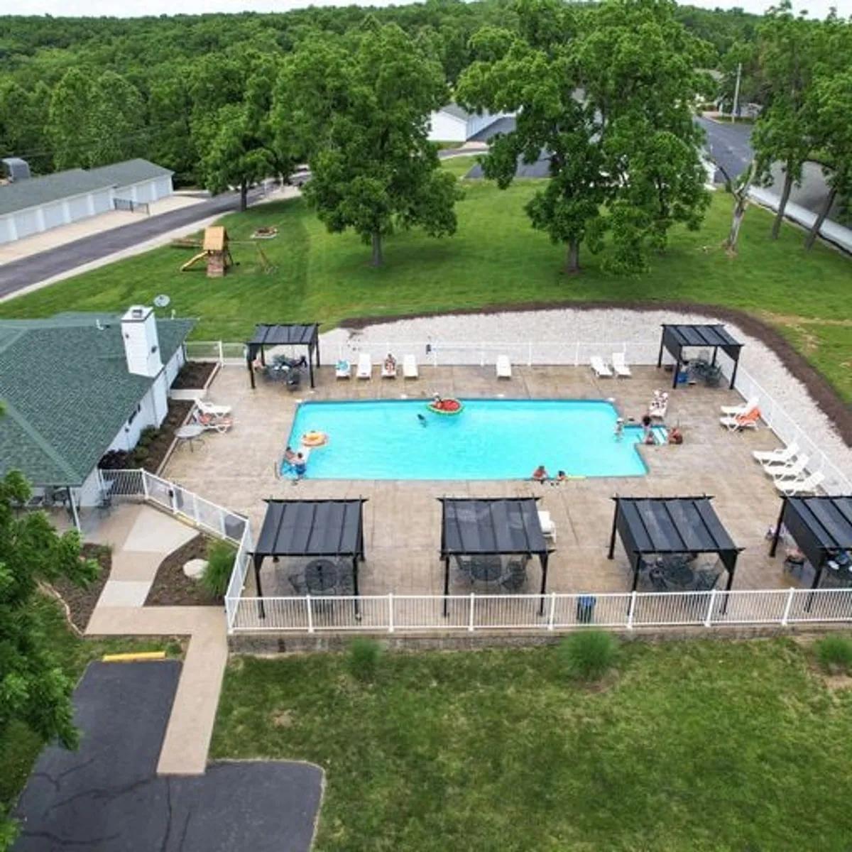 This image captures a vibrant outdoor swimming pool area surrounded by green lawns, gazeebo structures, and plenty of space for socializing. The scene evokes a relaxed mood ideal for group gatherings, sunbathing, or lakeside barbecues.
