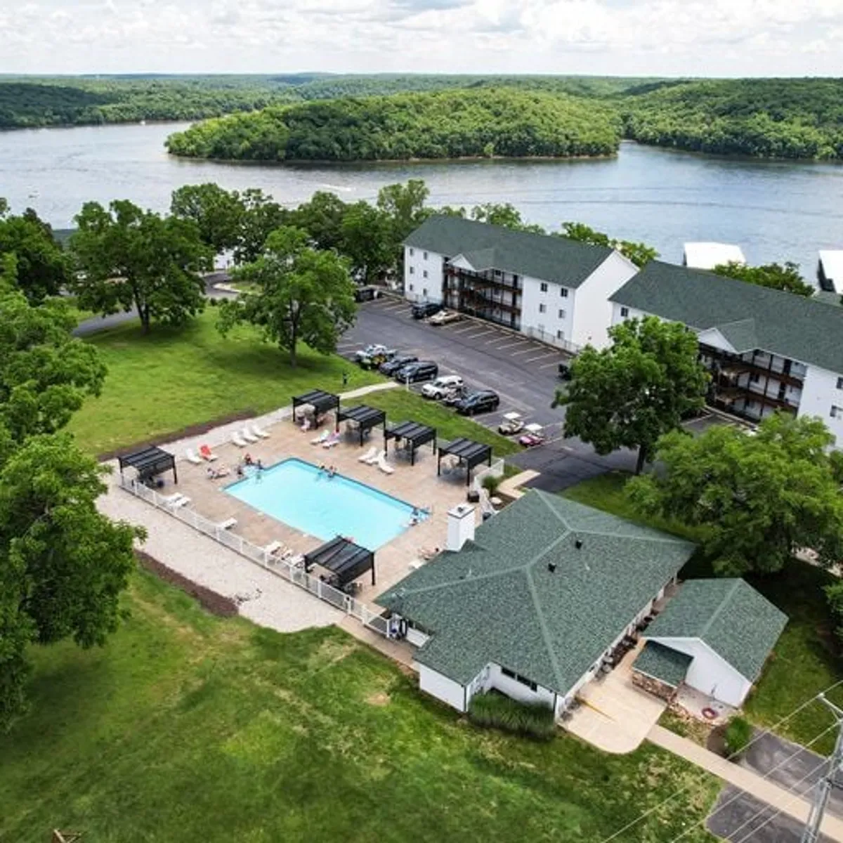 An inviting outdoor area featuring a swimming pool with lounge chairs, pavilions for shade, the lakeshore visible beyond, and adjacent buildings that suggest communal or social spaces. The scene exudes a relaxed mood, ideal for swimming, sunbathing, or casual gatherings by the water's edge.