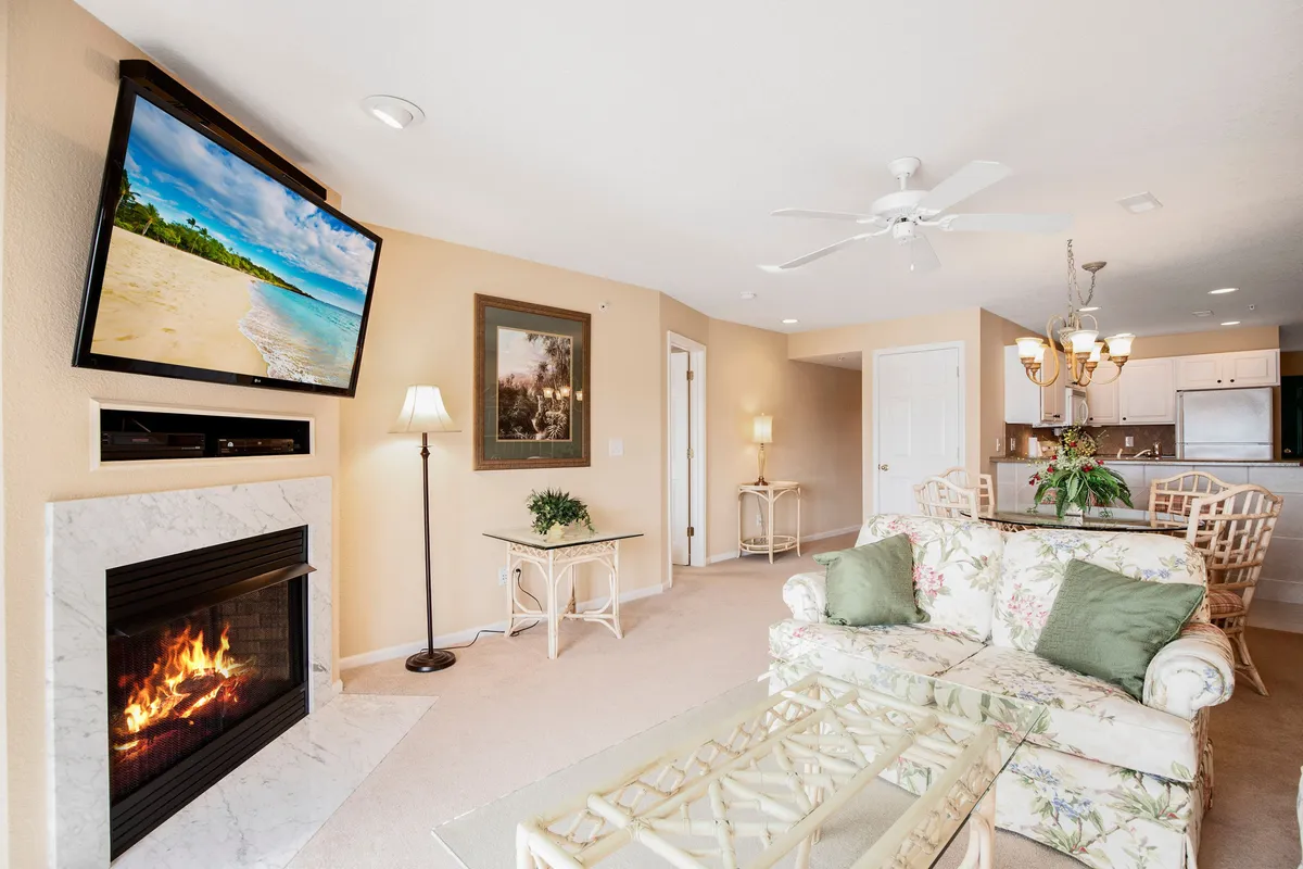 The living area features a floral-patterned sofa in front of a fireplace and a TV with a scenic beach image. The room is bright and welcoming, ideal for group gatherings or using the sofa sleeper for additional guests.