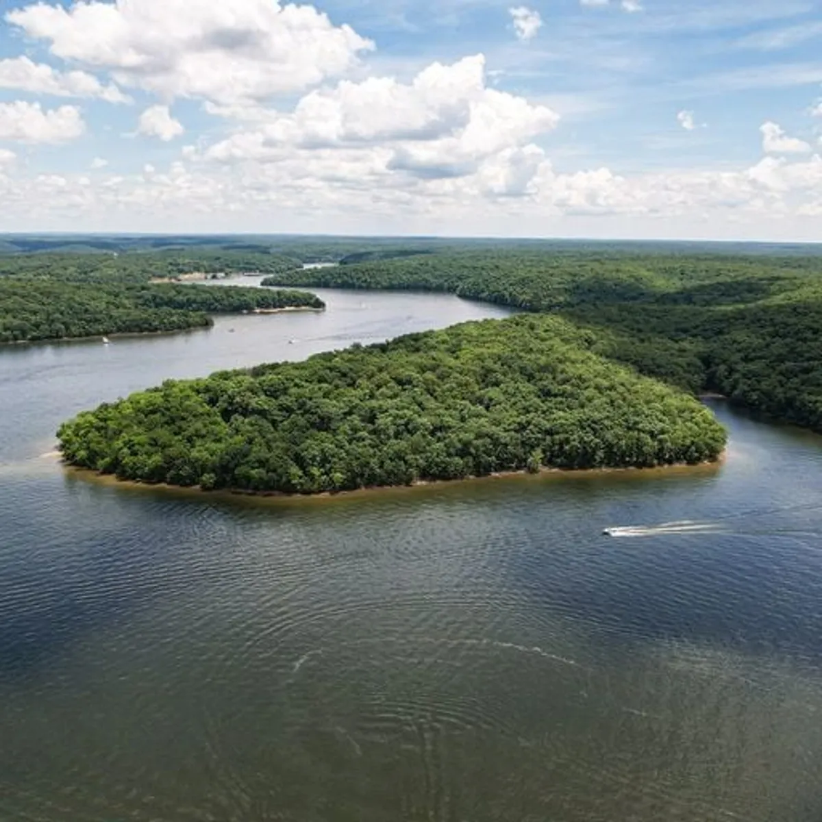 A sweeping view over a lush green island surrounded by calm, reflective water and distant tree-lined hills. The untouched landscape signals opportunities for hiking, wildlife spotting, and peaceful nature escapes.
