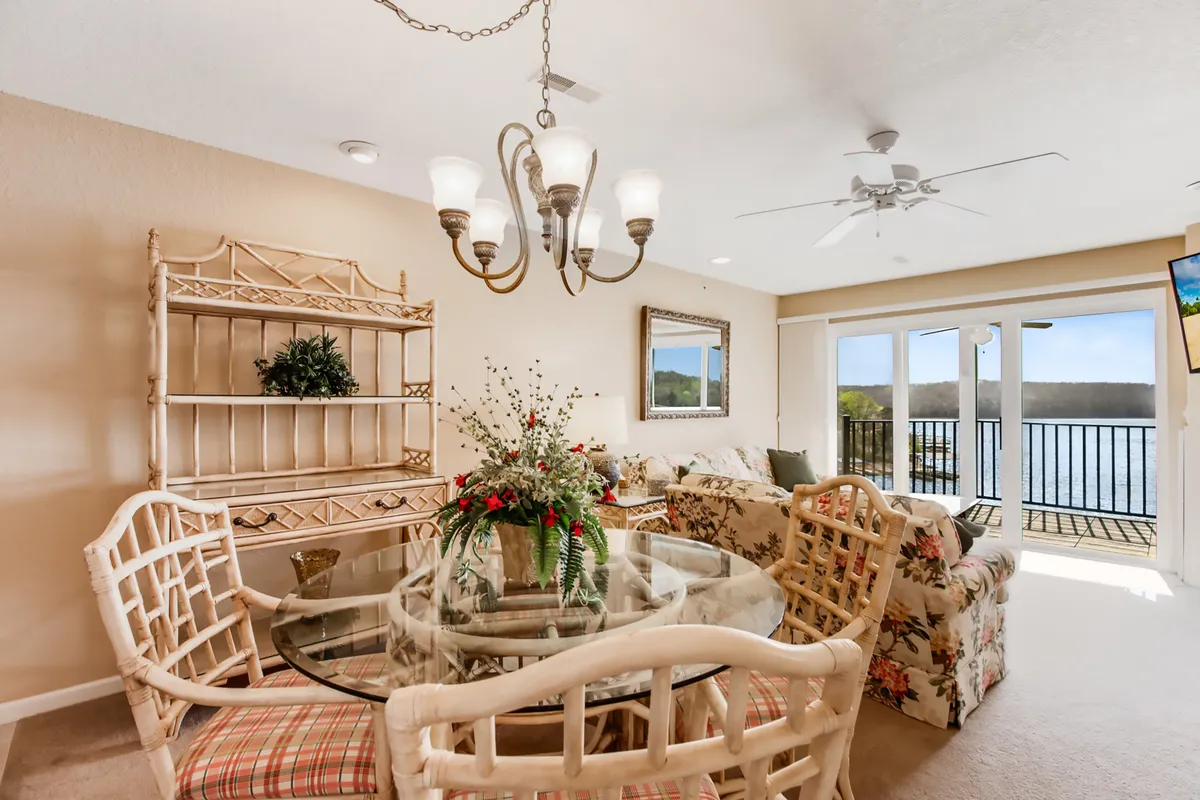 An interior scene featuring a glass dining table set for family meals, a floral patterned couch, a chandelier above, and a decorative shelf with greenery. The space feels cozy, inviting, and ready for entertaining guests or sharing special occasions indoors.
